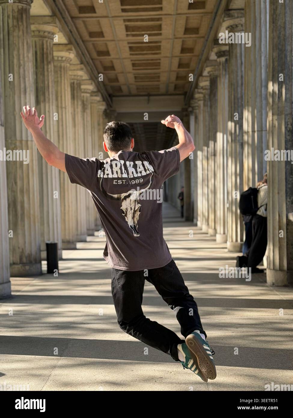 Young man walking with raised hands inside a historic stone colonnade in Berlin, Germany. Urban exploration, architectural perspective corridor view.⁠ - Smartphone Captured Stock Image