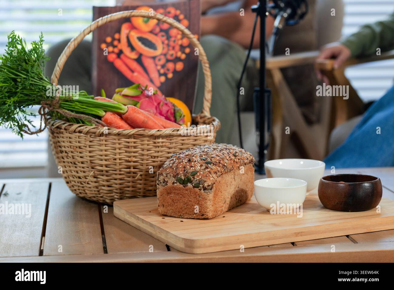 Seeded multigrain loaf resting on wooden cutting board on slatted table, wicker basket nearby Stock Photo