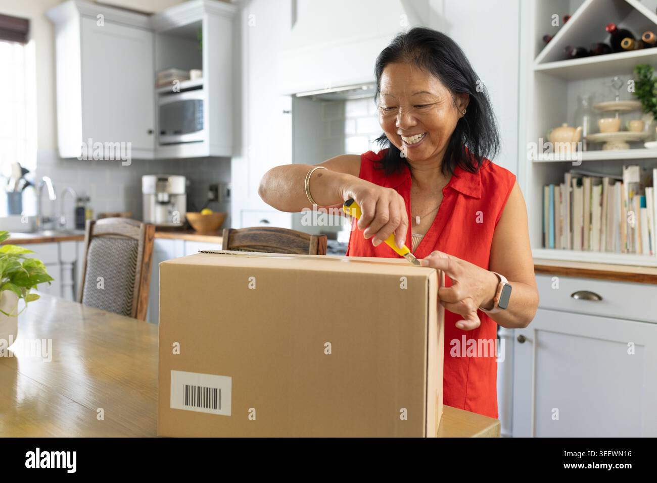 Senior Asian woman in red blouse standing at kitchen dining table opening box with yellow cutter Stock Photo