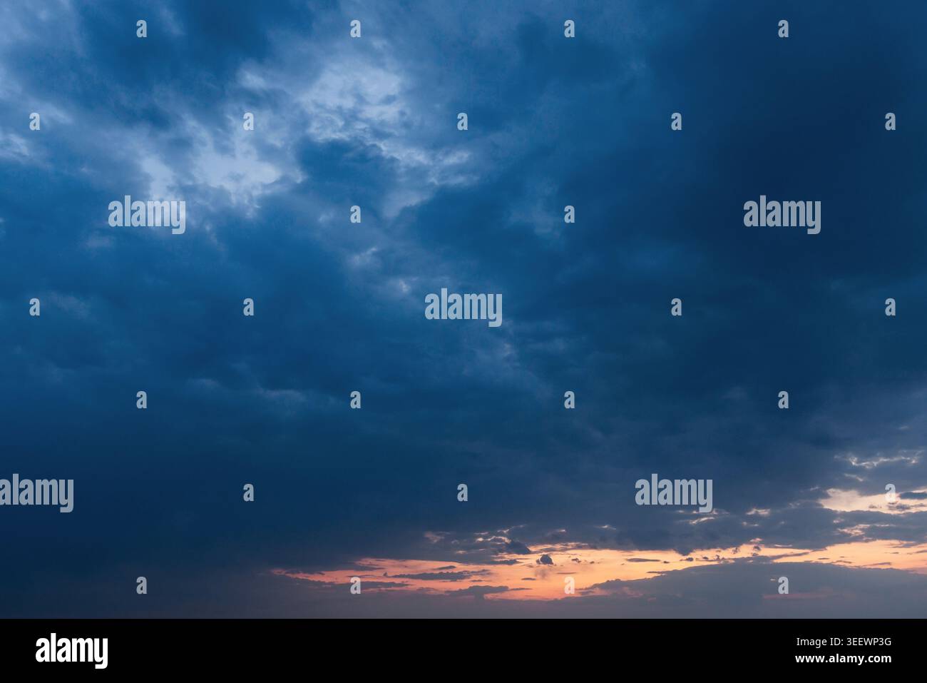 Dramatic blue storm clouds sweep across the evening sky above a thin orange sunset horizon, creating a moody, atmospheric scene ideal for weather, nat Stock Photo