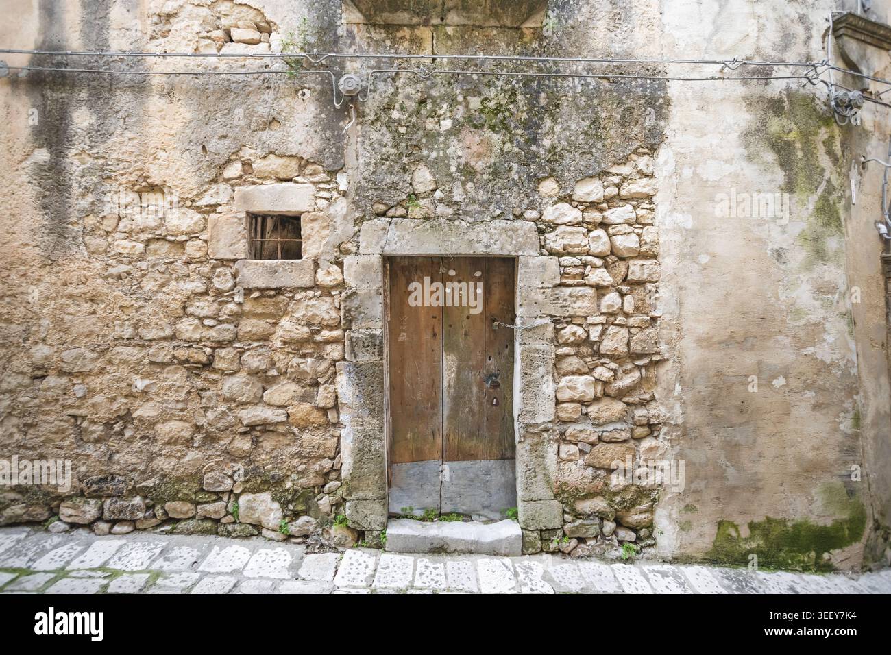 View of a rustic wooden door set in an ancient stone wall with a small barred window and mossy textures in Ragusa, Sicily, Italy. Stock Photo