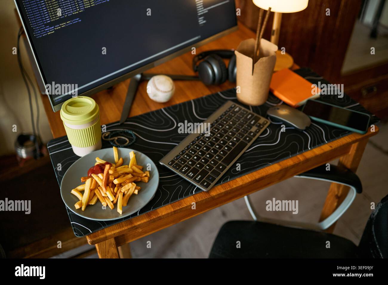 Close up of home office desk showing coding monitor, keyboard, smartphone, coffee cup, fries and headphones, illustrating remote work routine. Useful for productivity or work life balance Stock Photo