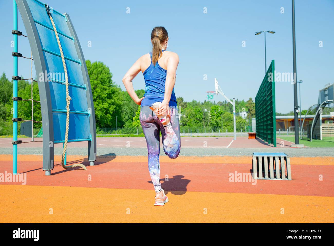 Athletic woman performs a quadriceps stretch on a sunlit urban sports ground. Mindful fitness routine for joyful longevity. Stock Photo