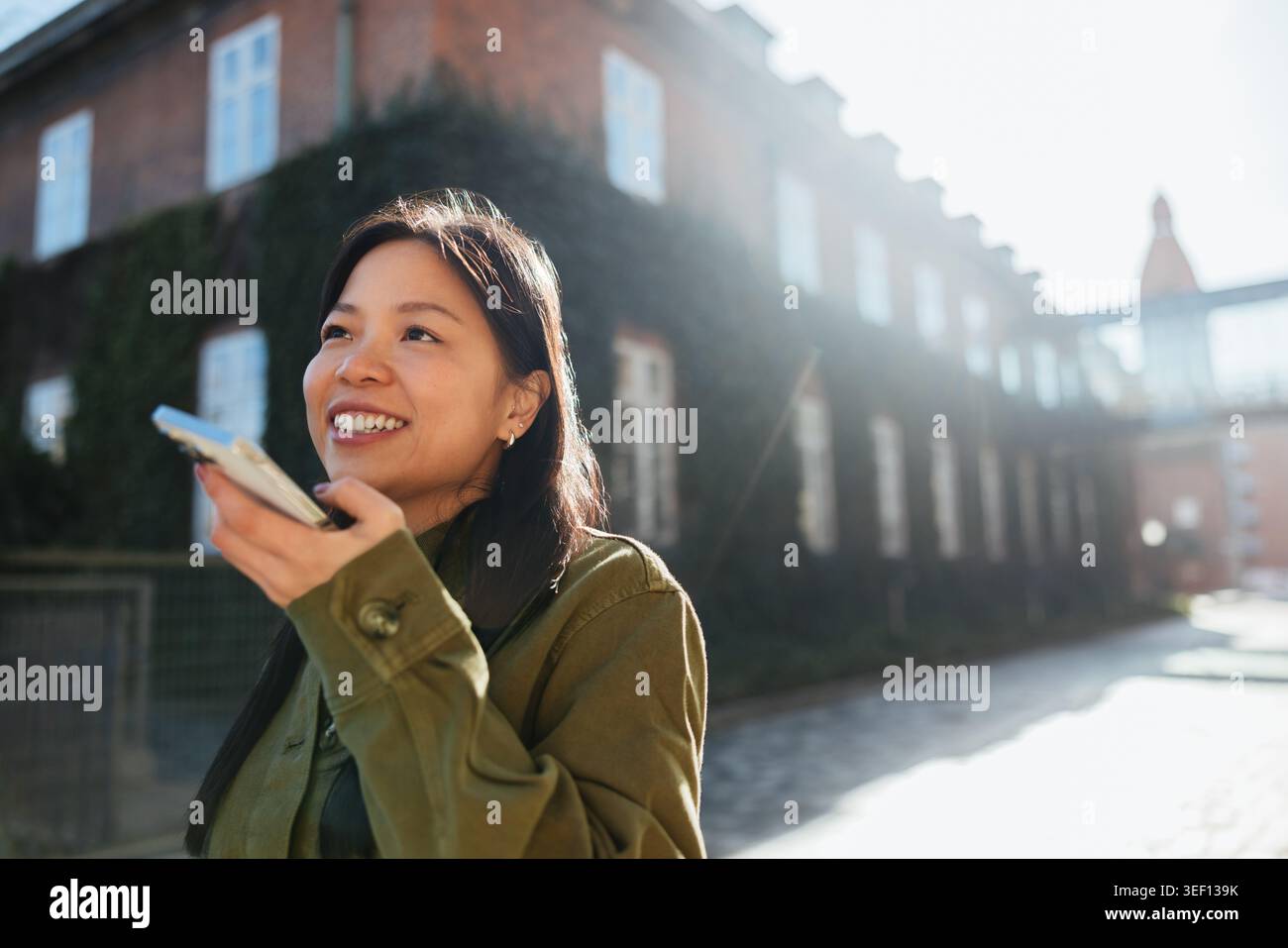 Smiling young Asian woman commuting through city streets, speaking into her phone to record a voice message while walking past historic brick building Stock Photo