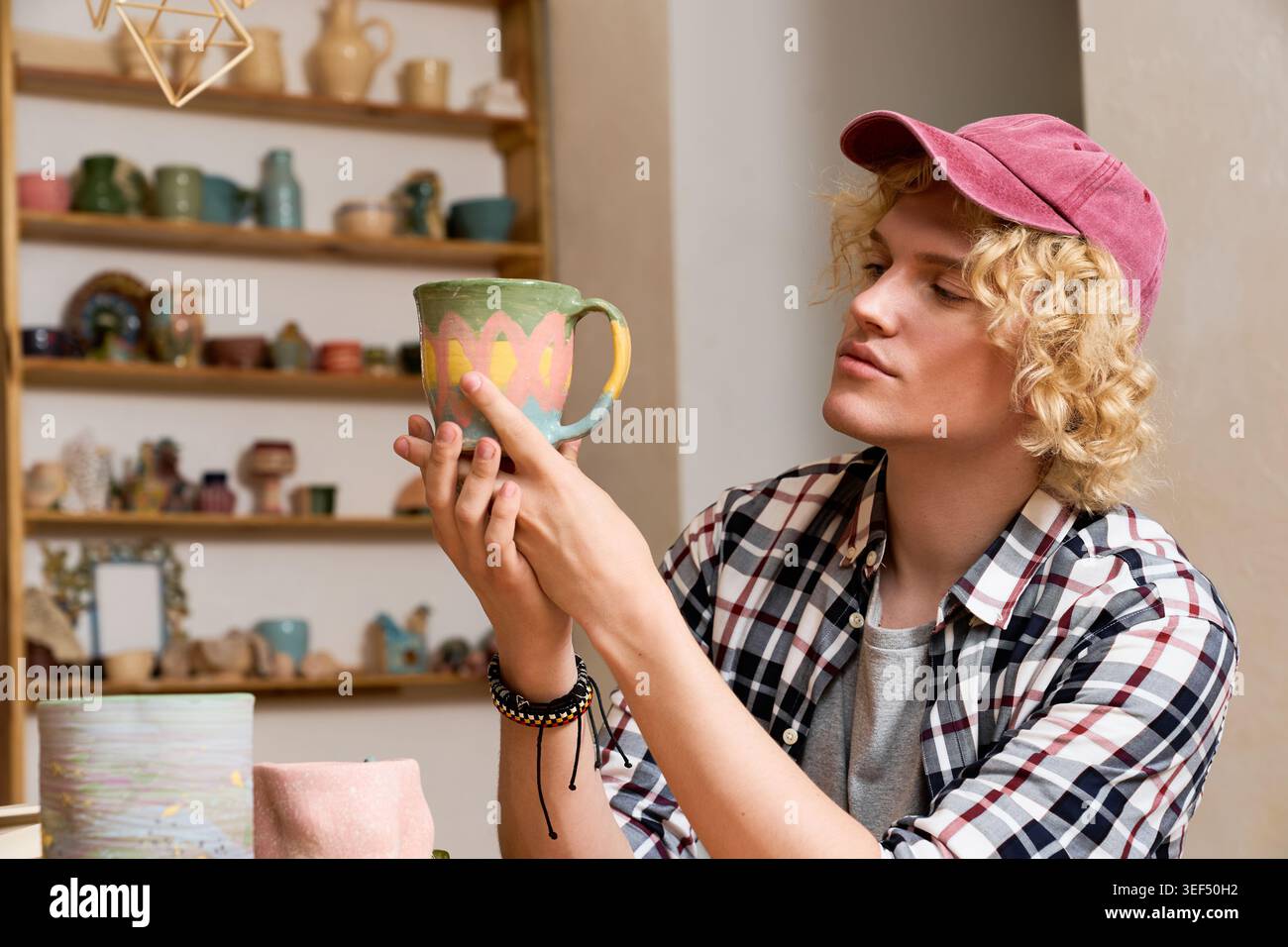 Young man carefully examines a colorful ceramic mug in his pottery workshop, showcasing skill Stock Photo