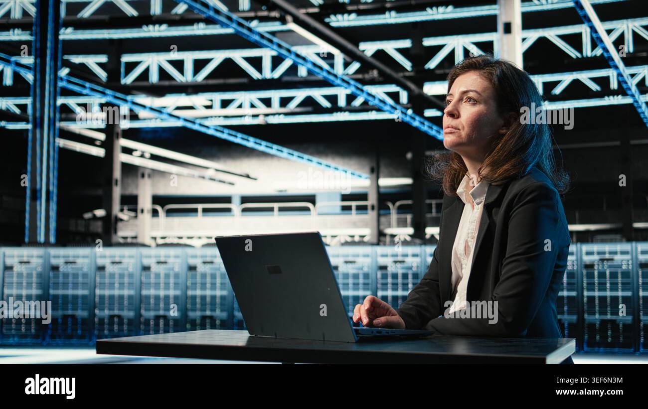 Data center software developer optimizing server load balancing using laptop. Woman in server farm at desk adjusting system parameters on notebook to improve efficiency Stock Photo