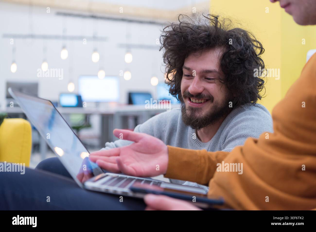 A candid close-up of a young man with a beard and curly hair laughing while looking at a laptop screen. The image captures a positive and high-energy Stock Photo