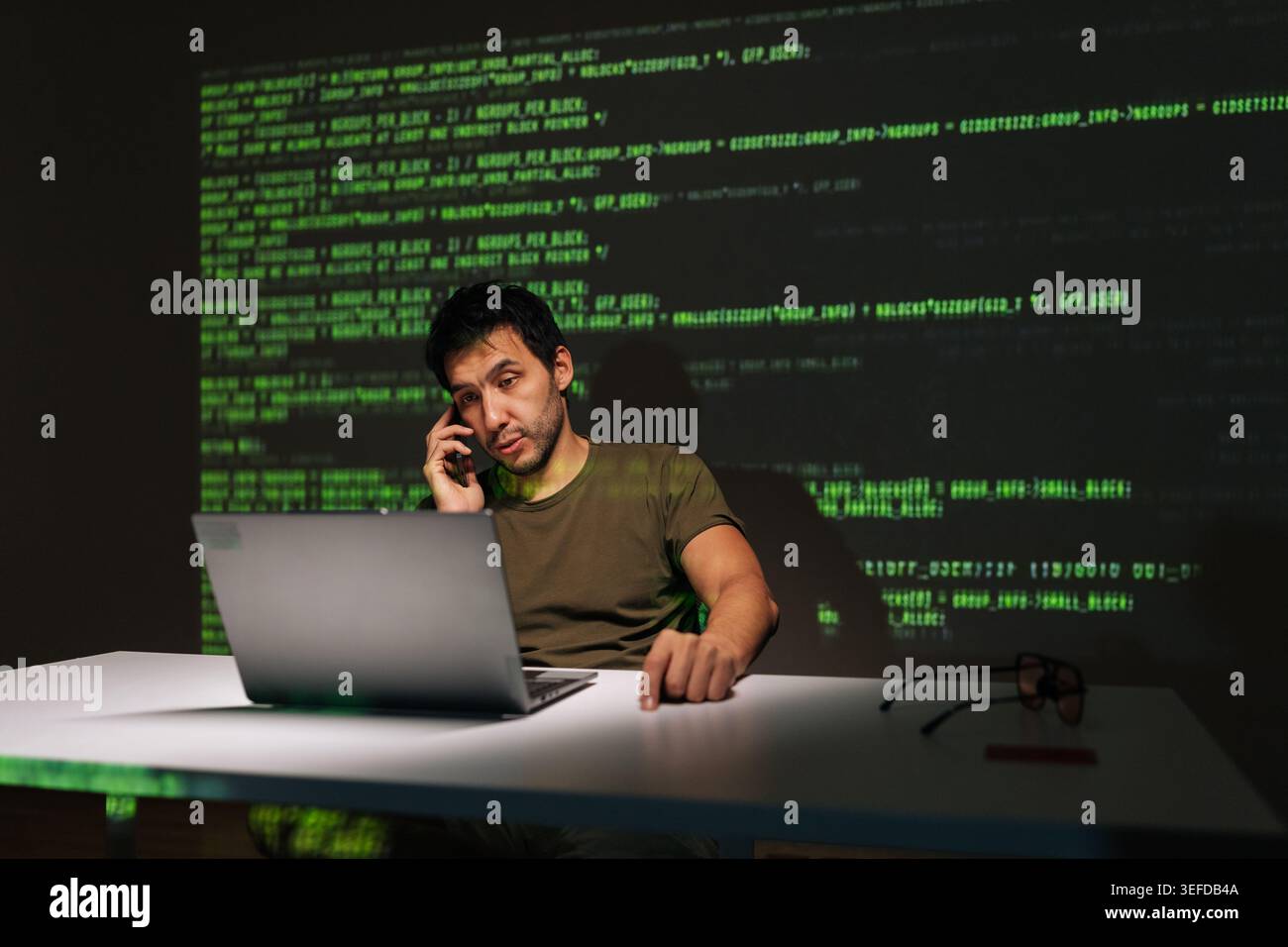 Programmer sitting at desk with laptop, deeply engaged in phone conversation while analyzing projected green code, symbolizing complex software Stock Photo