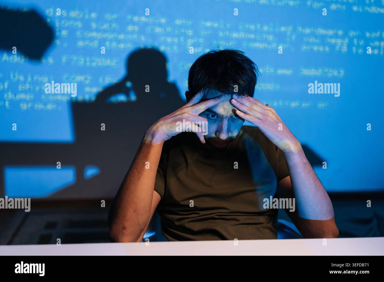 Stressed young male programmer facing challenging coding problem, feeling frustrated while working on laptop with lines of blue code projecting on Stock Photo