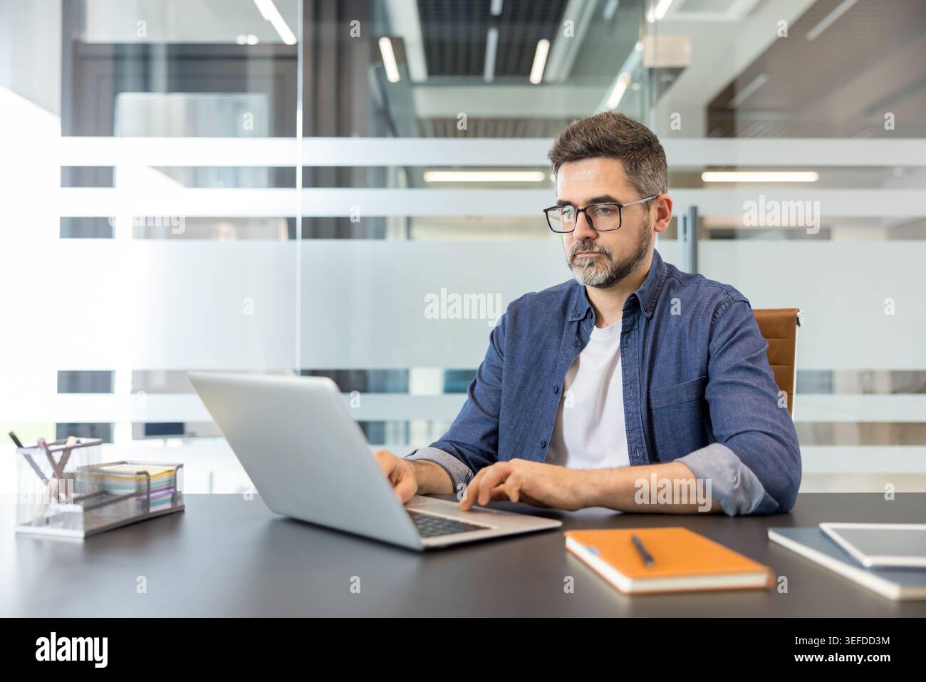 Focused man wearing glasses and a denim shirt sitting at a modern office desk, diligently typing on his laptop, embodying productivity and professional work environment Stock Photo