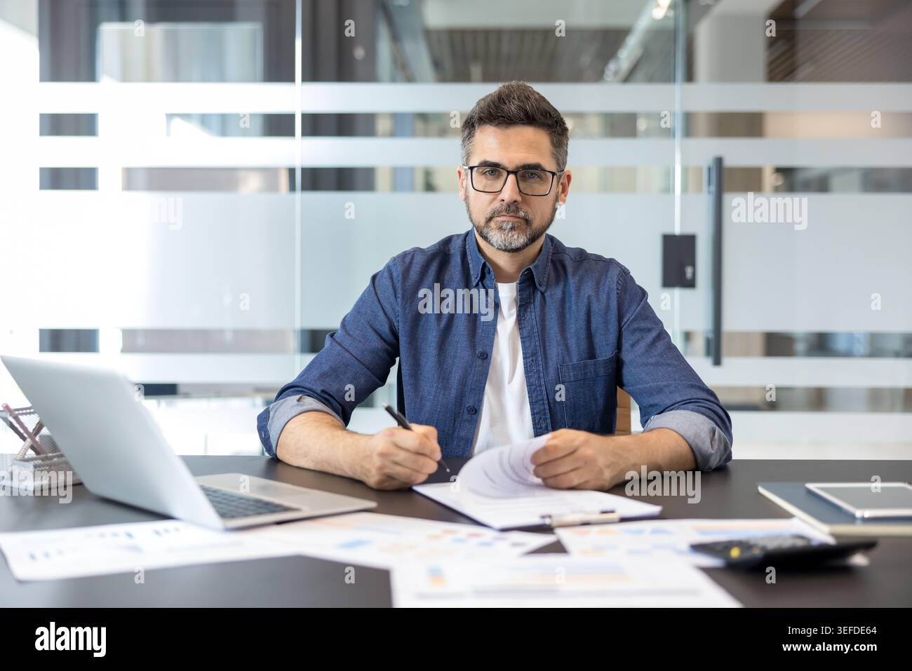 Businessman in a modern office writing notes and reviewing financial reports, representing professionalism, strategy, finance, and successful business operations Stock Photo