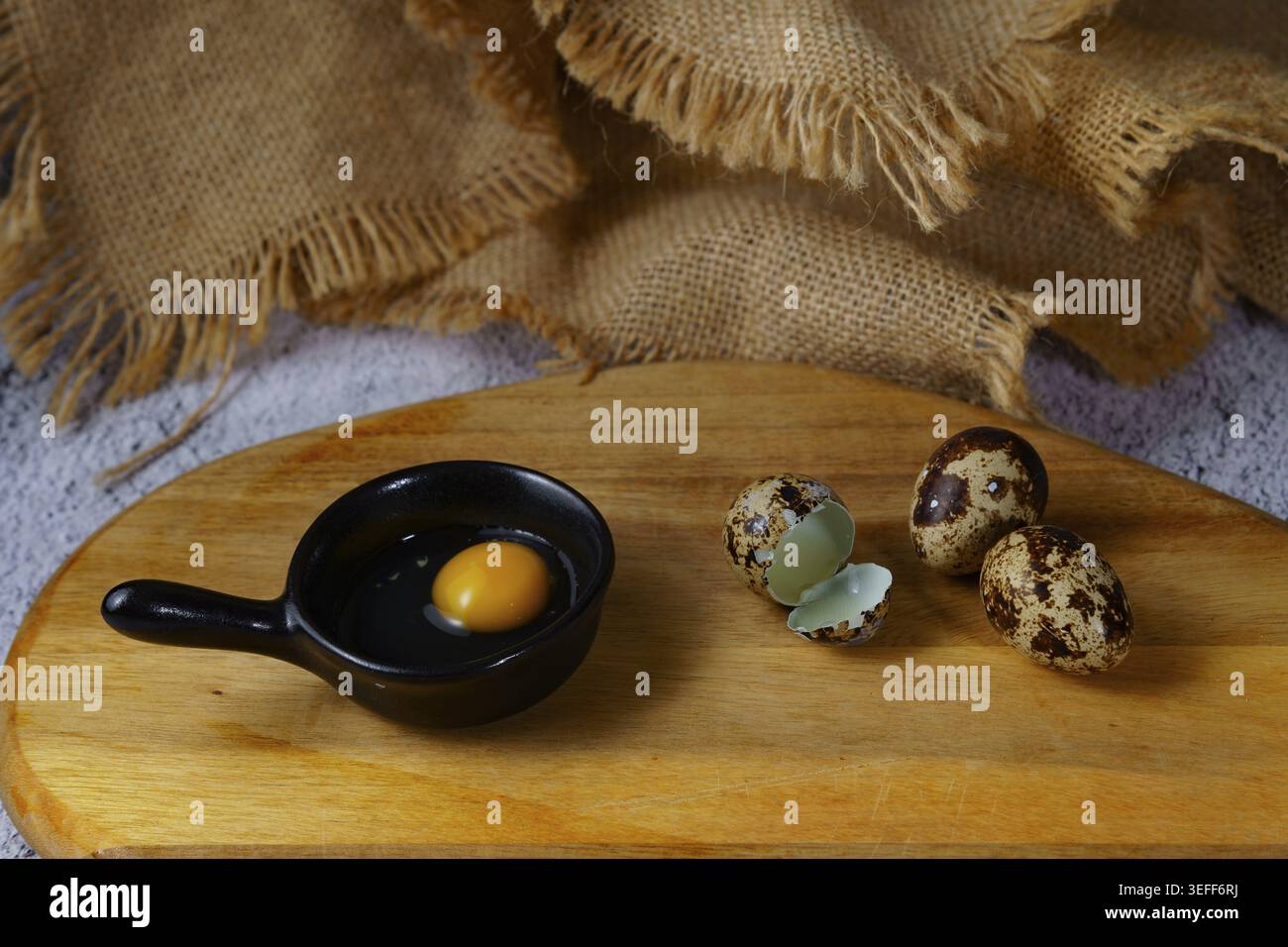 Quail egg yolk inside a small black ceramic bowl, with broken shells and whole quail eggs on a rustic wooden cutting board, preparing ingredients Stock Photo