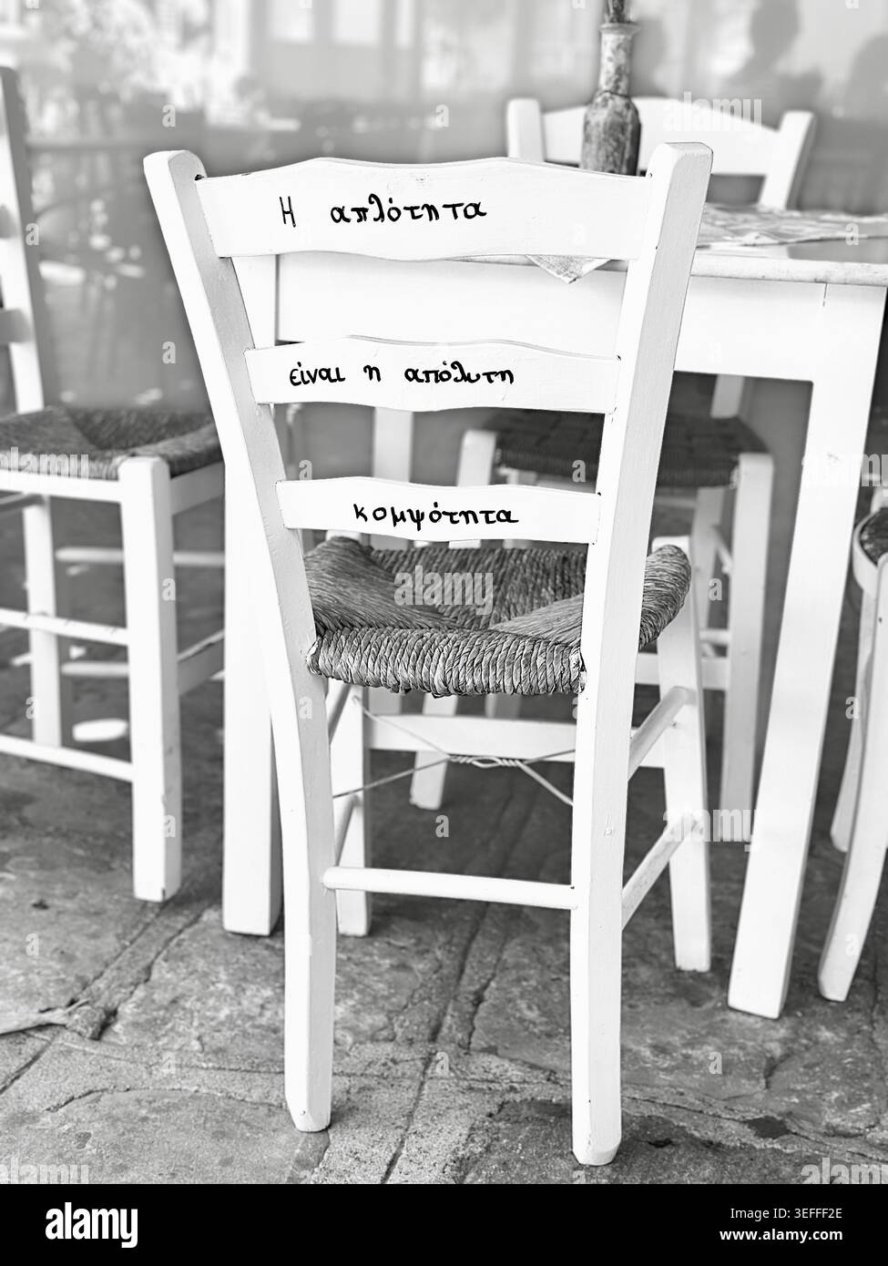 Black and white iphone photography of straw chair and wooden table with quote on the back in Greek 'simplicity is the ultimate elegance' - Smartphone Captured Stock Image