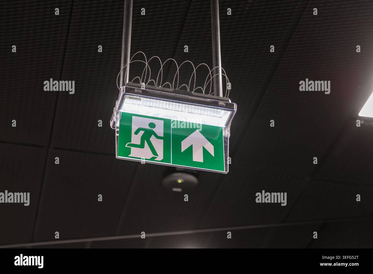 A bright glowing emergency exit sign with a directional arrow is mounted on a ceiling, representing safety regulations, clear navigation, disaster pre Stock Photo