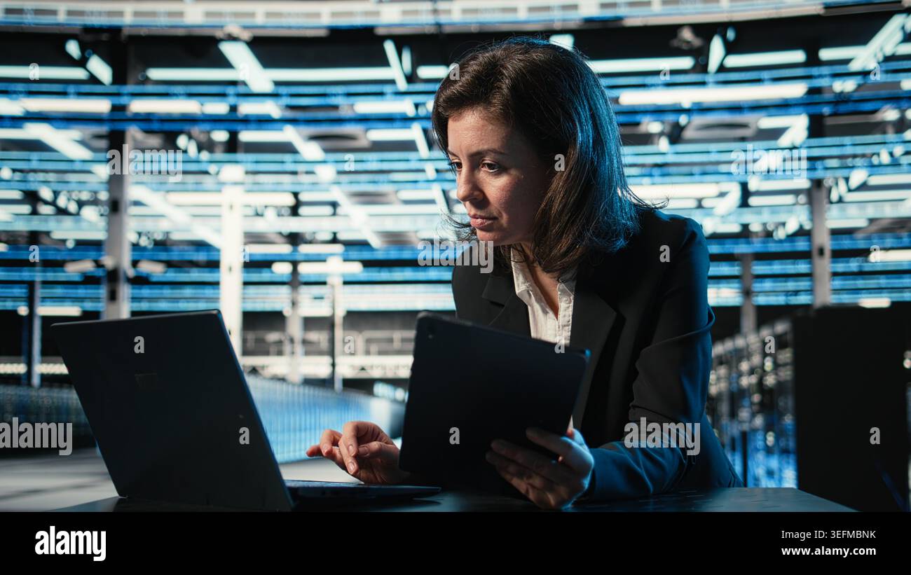 Data center software developer optimizing equipment rigs using devices. Woman in server farm running programming script on laptop and tablet, adjusting system parameters Stock Photo