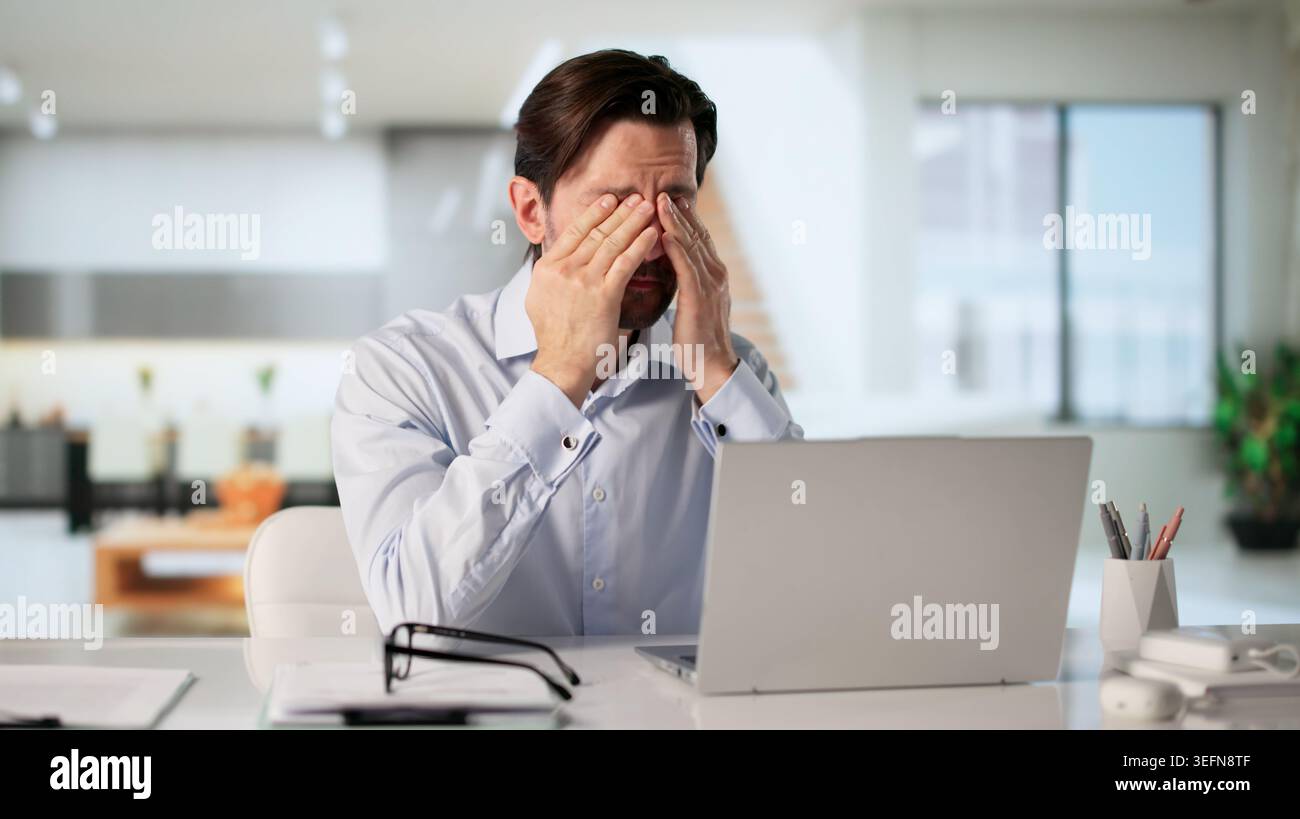 Tired Man Rubbing Sore Eye While Working On Laptop Feeling Stressed. Stock Photo