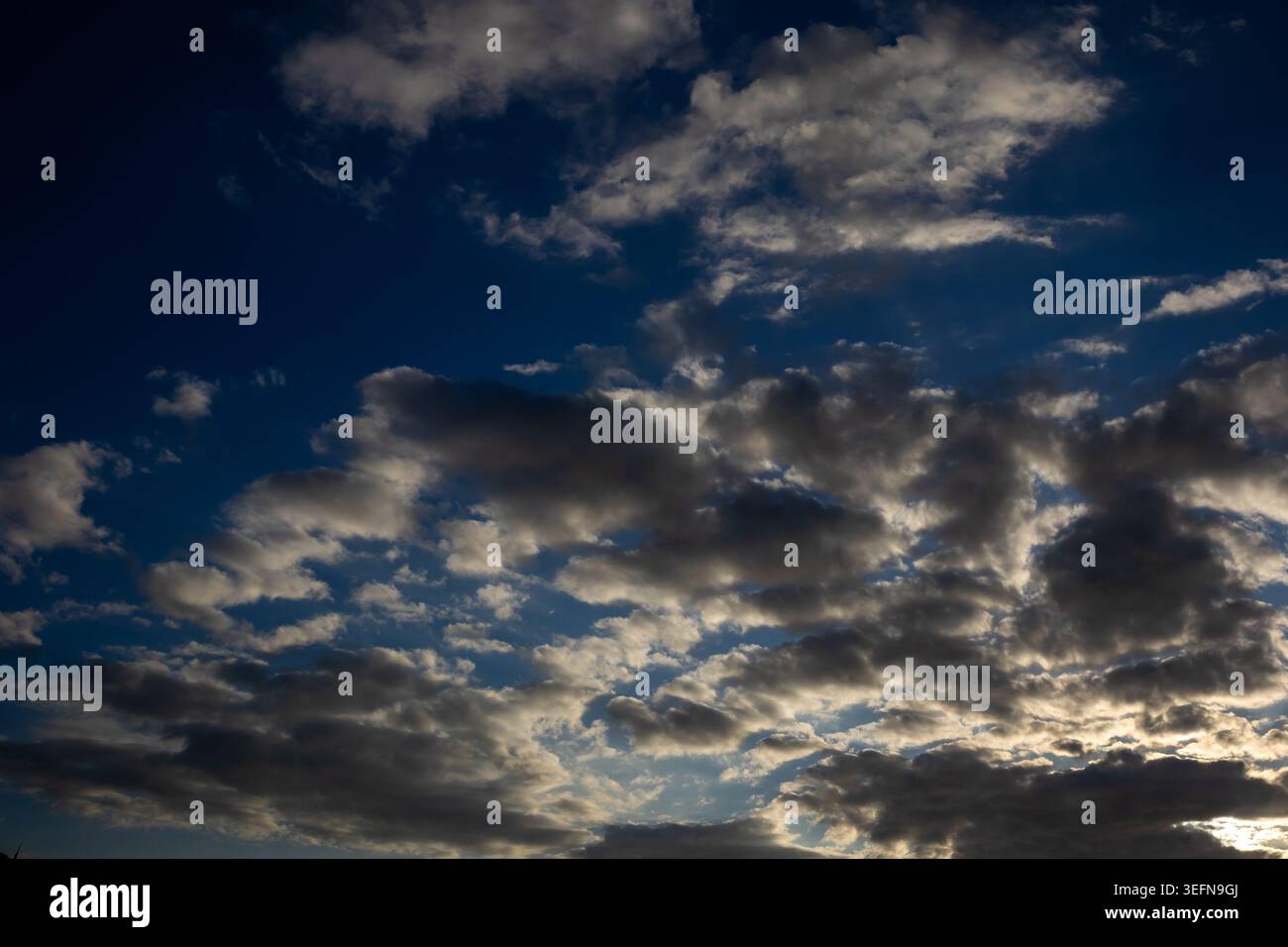 Broken cloud formations scattered across deep blue sky with warm light catching cloud bases from below Stock Photo