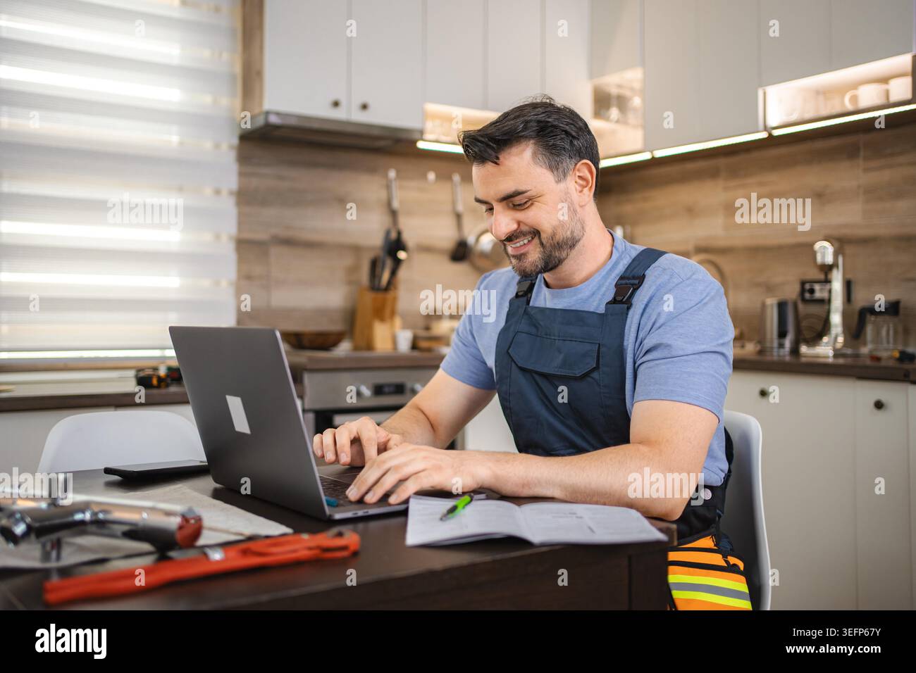 Smiling repairman working on laptop in kitchen with plumbing tools, using computer for online learning Stock Photo