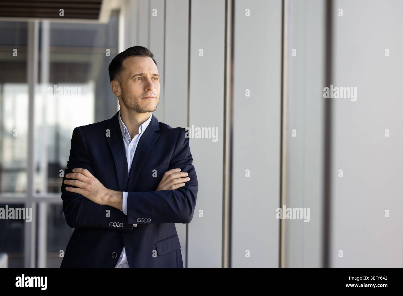 Thoughtful businessman executive standing by office window with folded arms Stock Photo