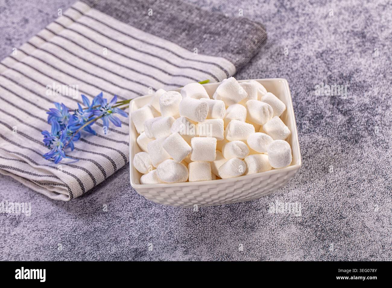Sweet white mini marshmallows fill decorative bowl, set beside striped kitchen towel delicate blue flowers. Light grey textured background provides cl Stock Photo
