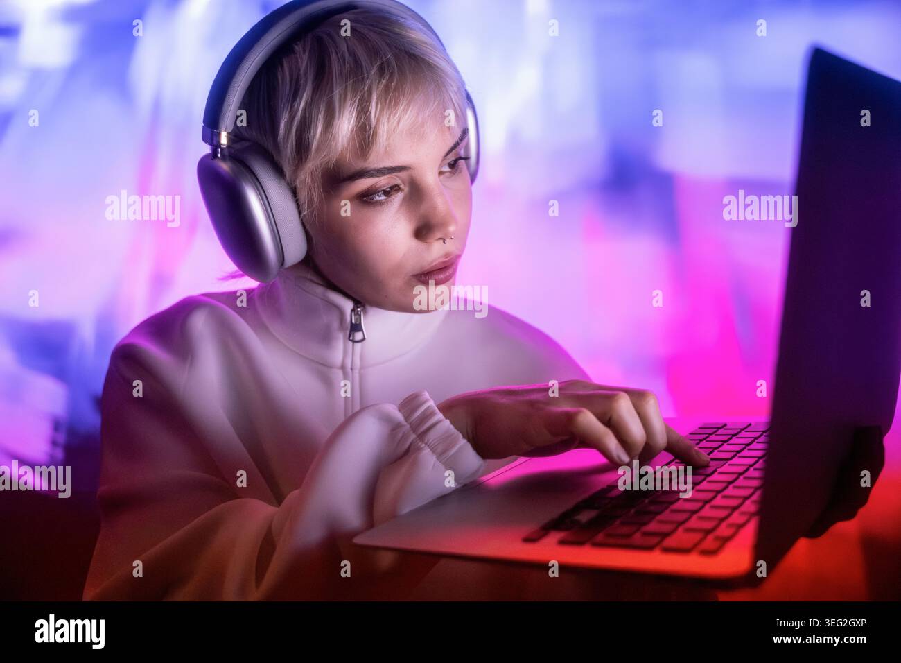 Futuristic woman wearing headphones and a white jacket works on a laptop, focused on typing in a neon pink and blue glow, creating a modern cyberpunk Stock Photo