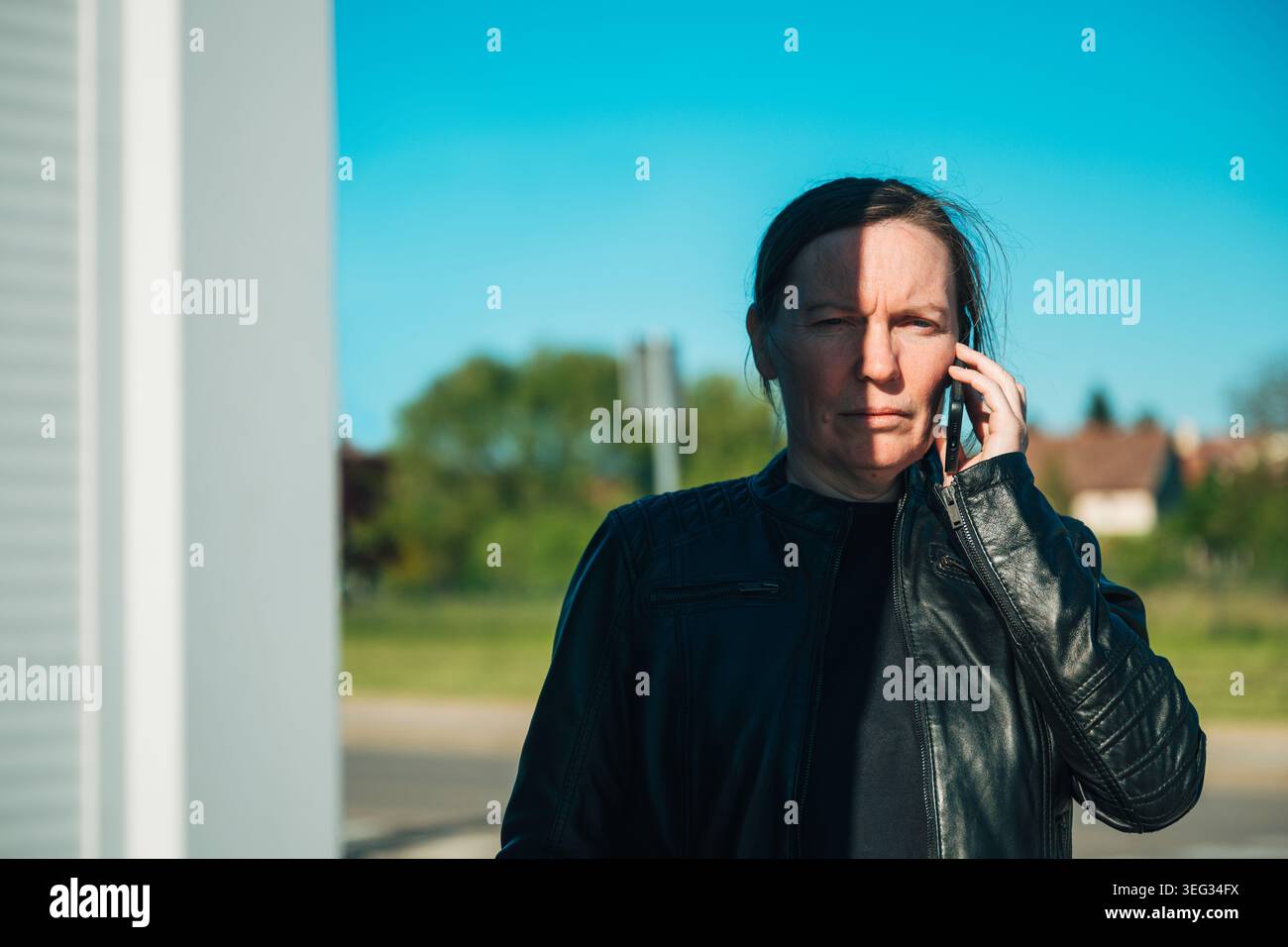 Serious middle-aged woman standing outdoors in a suburban area, wearing a black leather jacket while making a phone call on a mobile device under brig Stock Photo