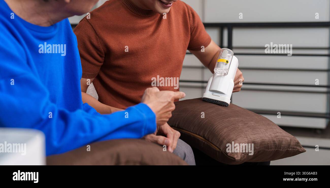 Asian son helping senior father with home cleaning using lint remover. Stock Photo