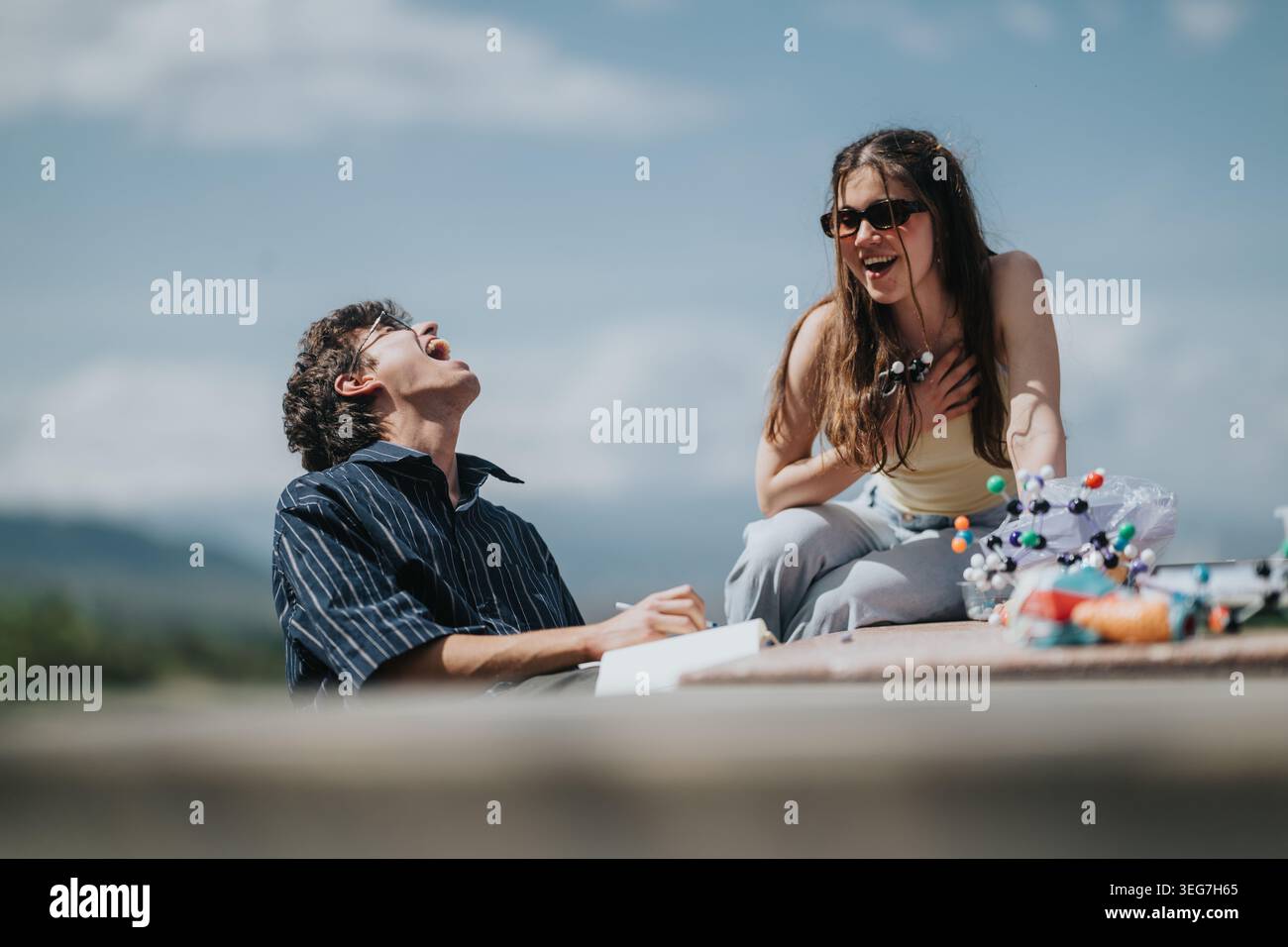 Two friends laughing outdoors while studying with a molecular model on a sunny day Stock Photo