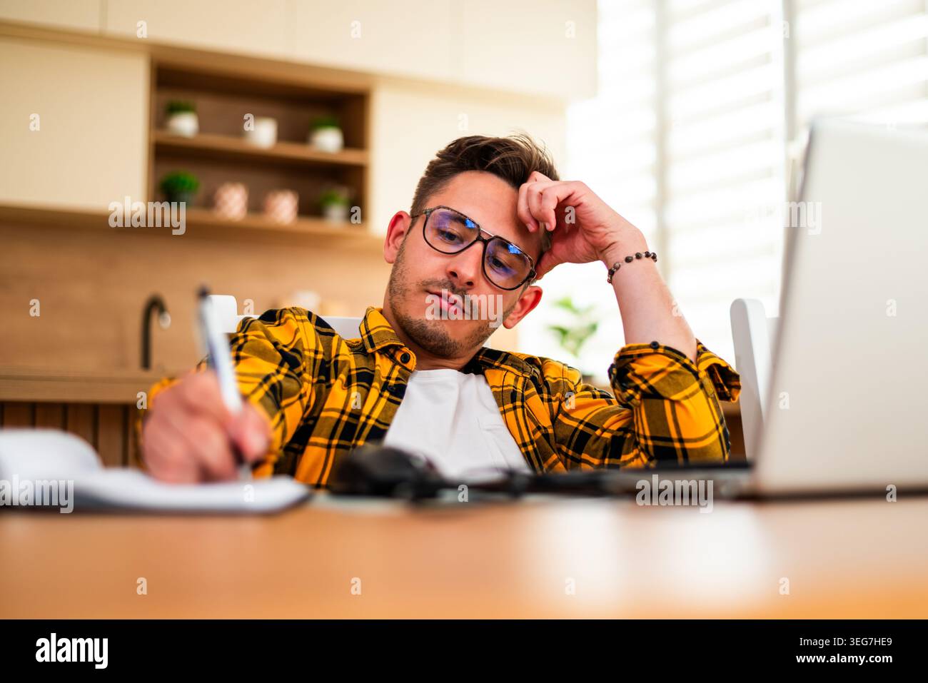 Man studies his laptop screen while writing in a notebook, suggesting pressure from work or study. Stock Photo