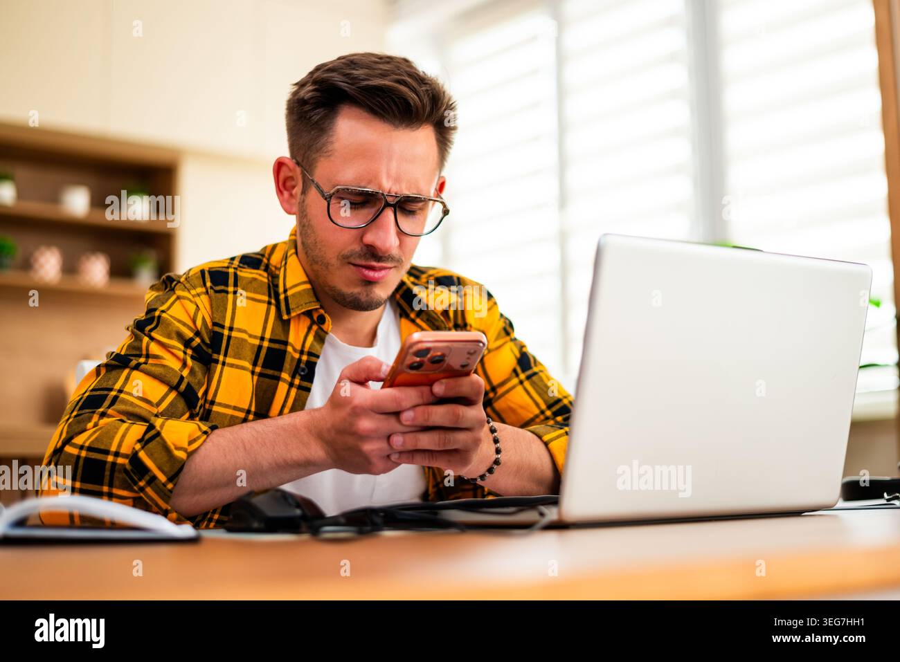 With a tense expression, a man scrolls through his phone at his desk, suggesting stress from work or unexpected news. Stock Photo