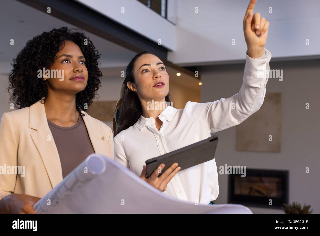 Diverse female coworkers holding blueprints and tablet, pointing and inspecting modern mezzanine Stock Photo