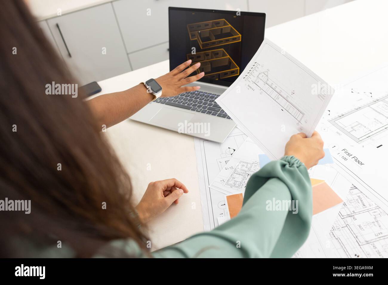 Diverse female coworkers reviewing blueprints on white table, pointing at laptop 3D wireframe Stock Photo