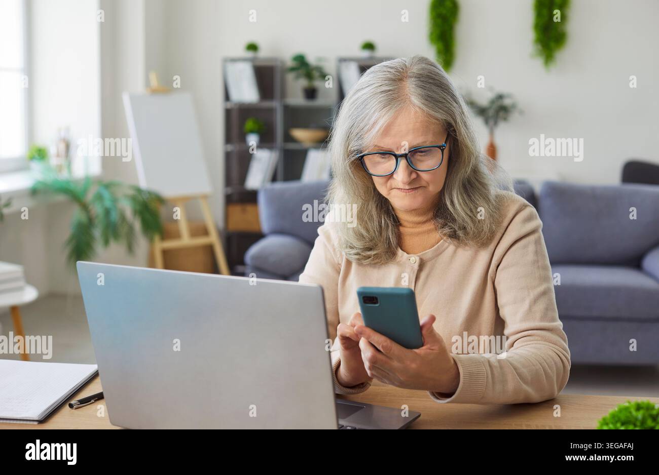 Old senior woman sitting at table holding smartphone looking down at screen, interacting online Stock Photo