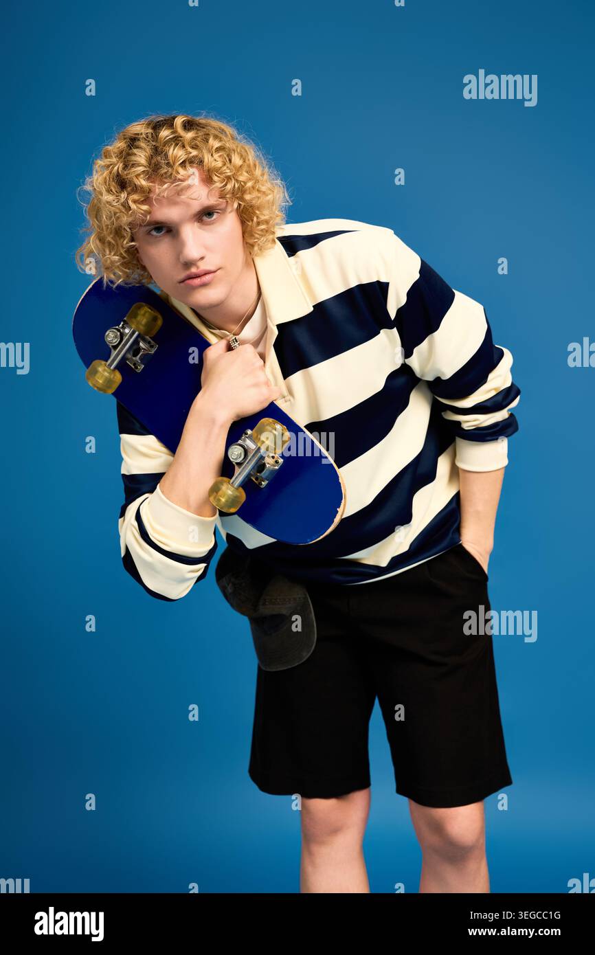 Blonde, curly haired student showcases style while holding a skateboard in a lively studio. Stock Photo