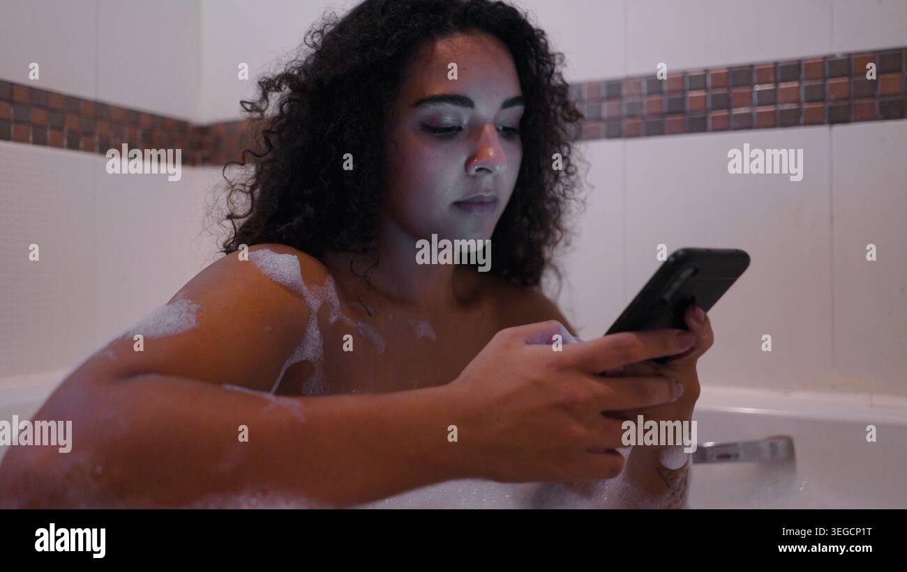 Young woman relaxing in bubble bath while connecting with smartphone Stock Photo