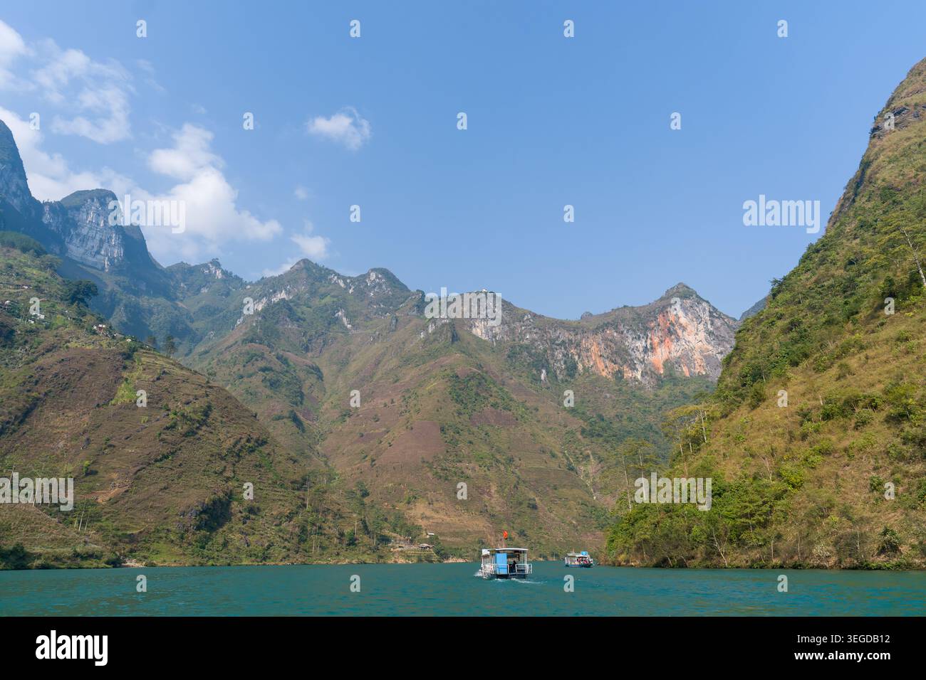 Serene lake surrounded by towering green mountains. Small boats drift on calm, turquoise waters. Clear blue sky with soft white clouds above. Rugged c Stock Photo