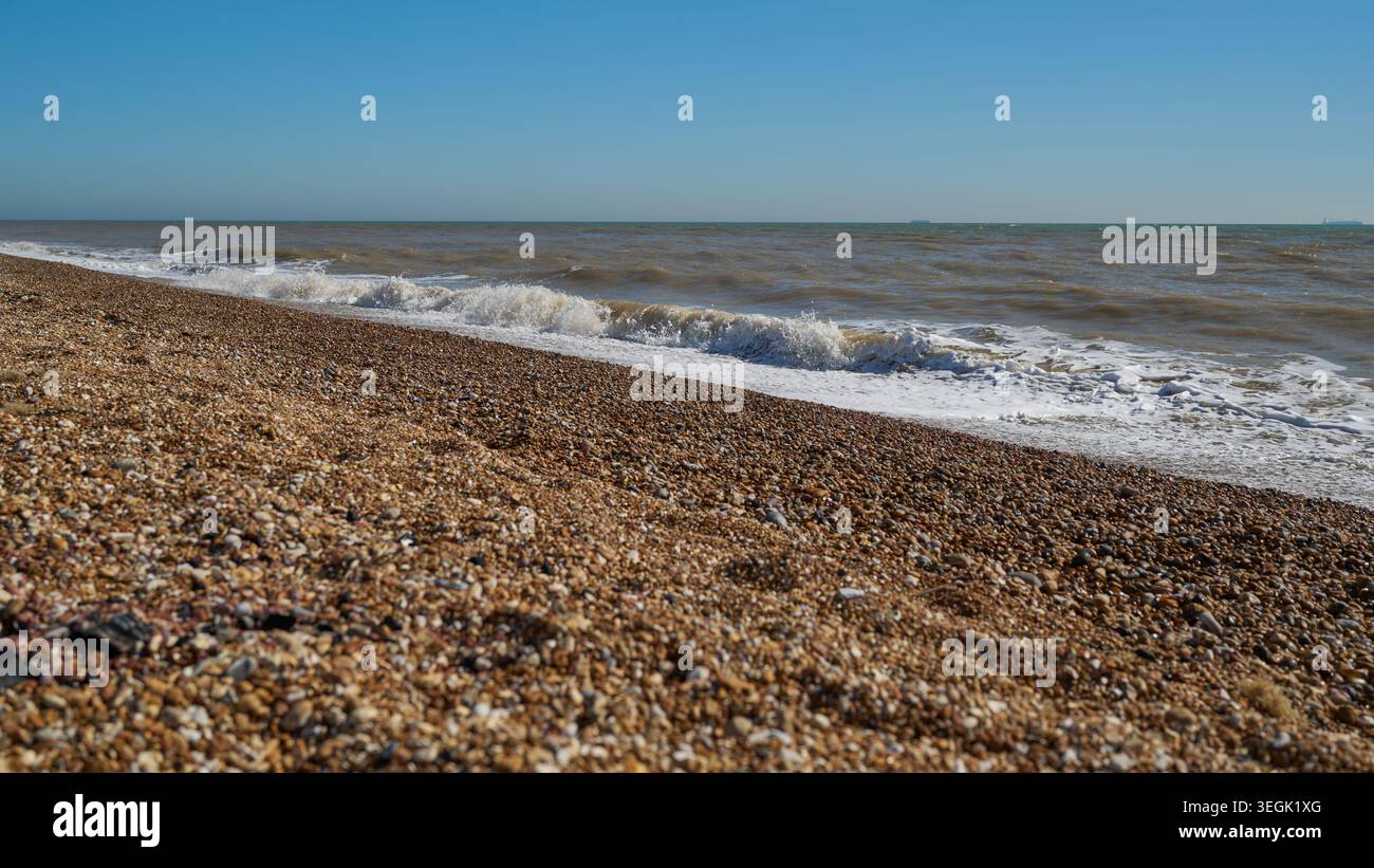 Pebble beach shoreline with incoming waves under clear blue sky. Stock Photo