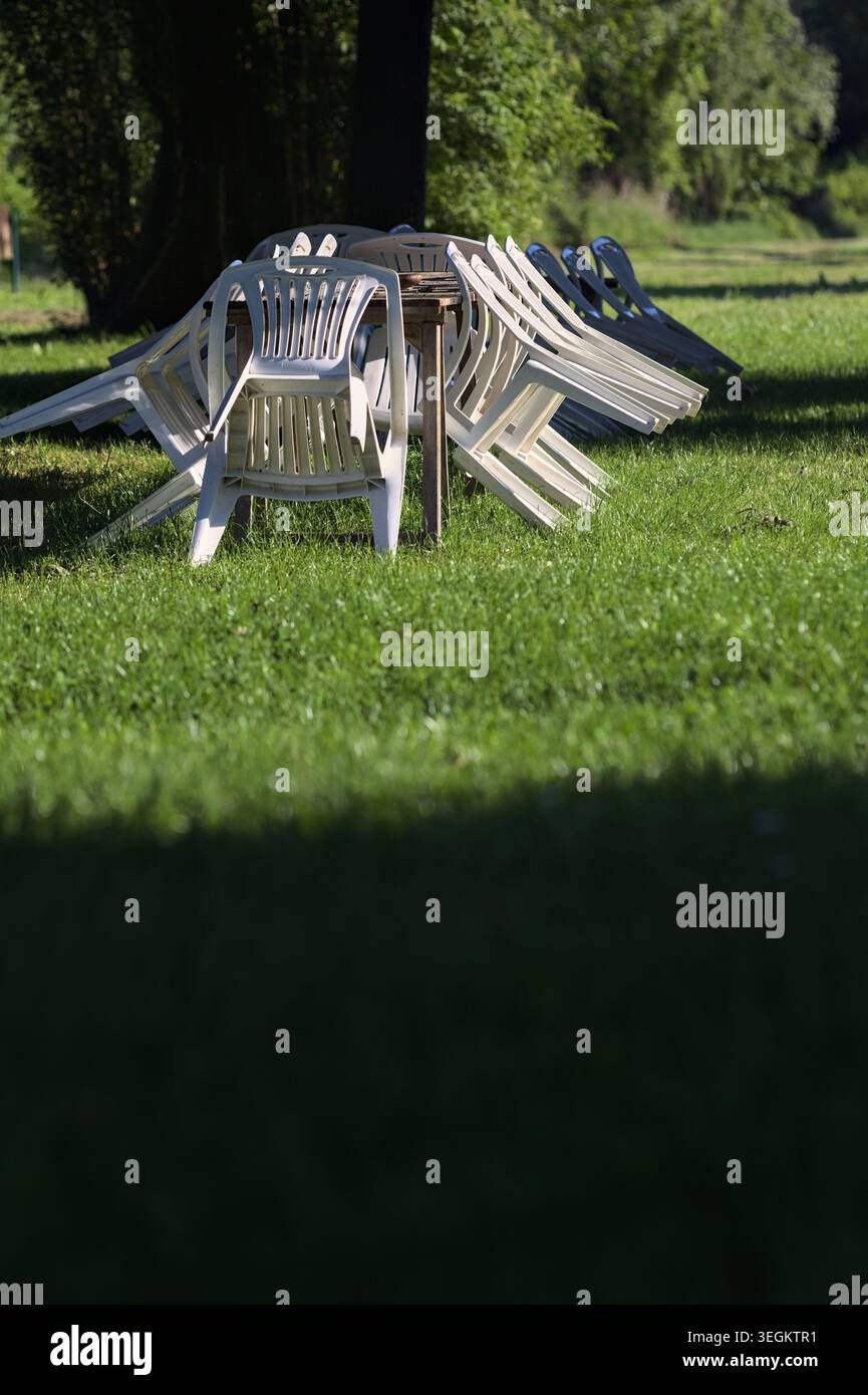 Plastic chairs leaning on a wooden table in the middle of a lawn, in a park, at sunset, Stock Photo