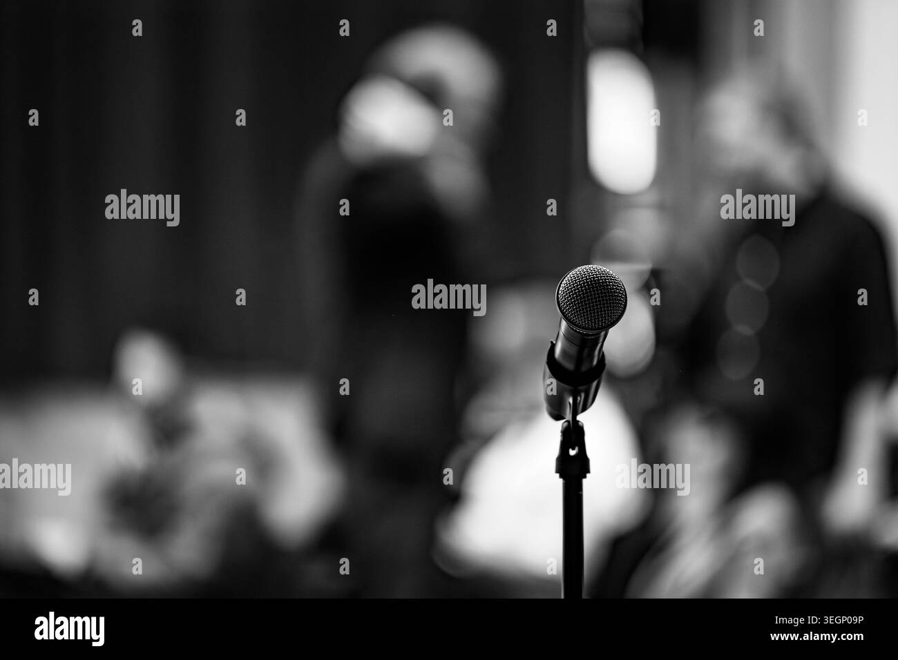 A vocal microphone stands on a mount in a dimly lit venue with blurred figures of people sitting in the background during an indoor public event or pr Stock Photo
