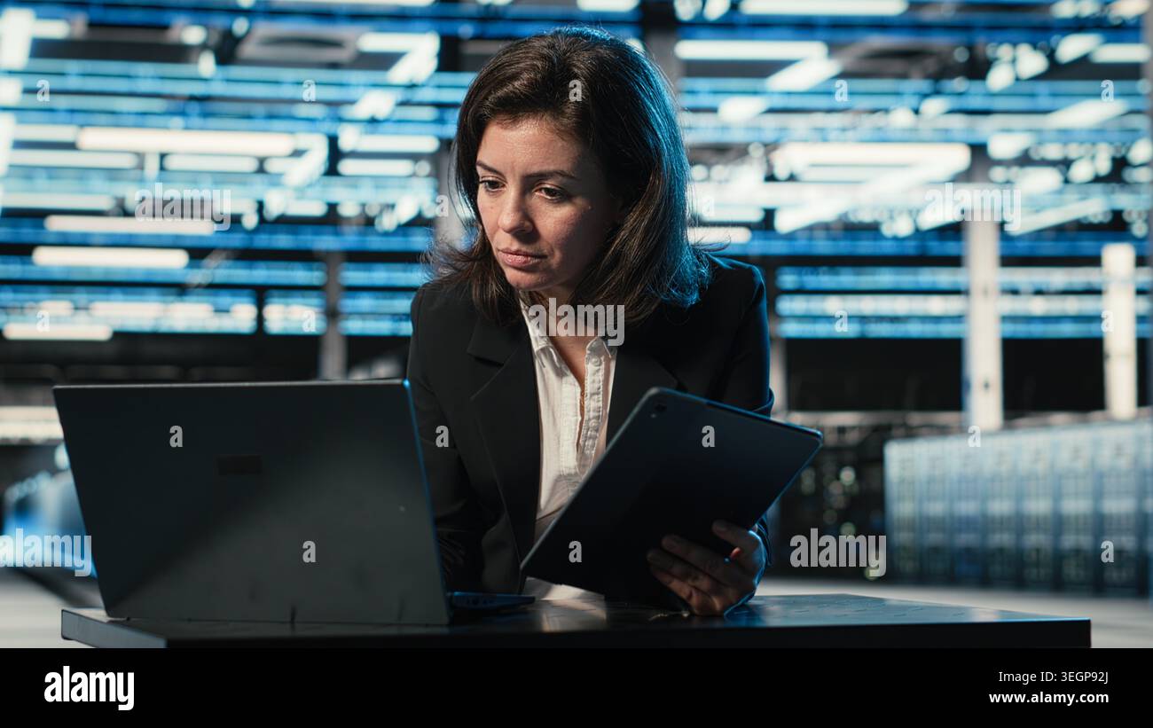 Data center software developer optimizing equipment rigs using devices. Woman in server farm running programming script on laptop and tablet, adjusting system parameters Stock Photo