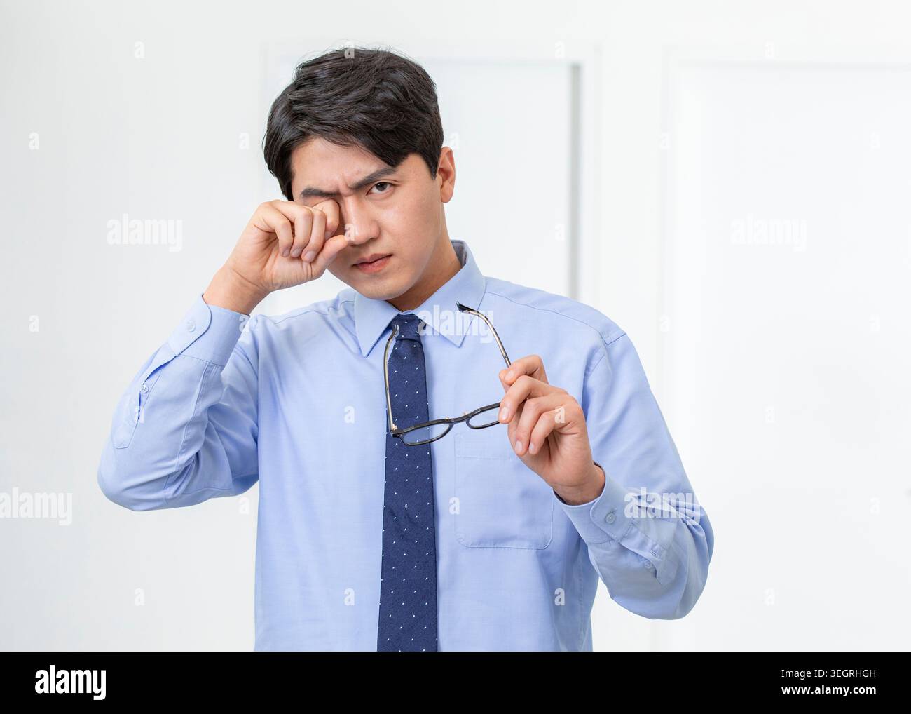A young Asian man in a white shirt and blue tie rubs his tired eyes in a modern office setting. Stock Photo