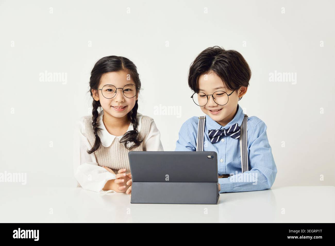 Two children in glasses sit at a table, using a tablet. Stock Photo