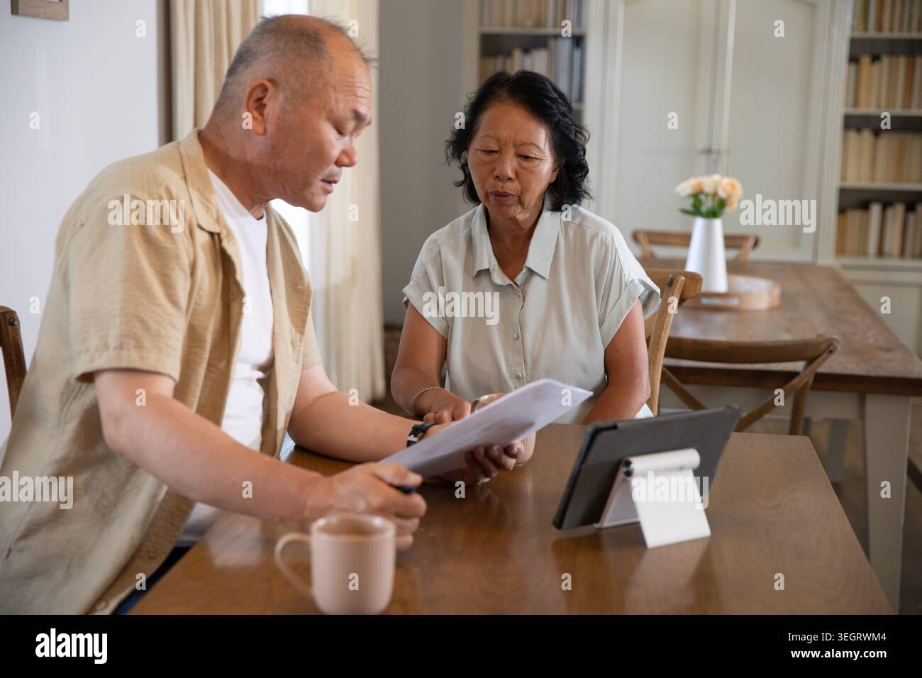 Asian couple examining printed documents at home dining table while using tablet and coffee mug Stock Photo