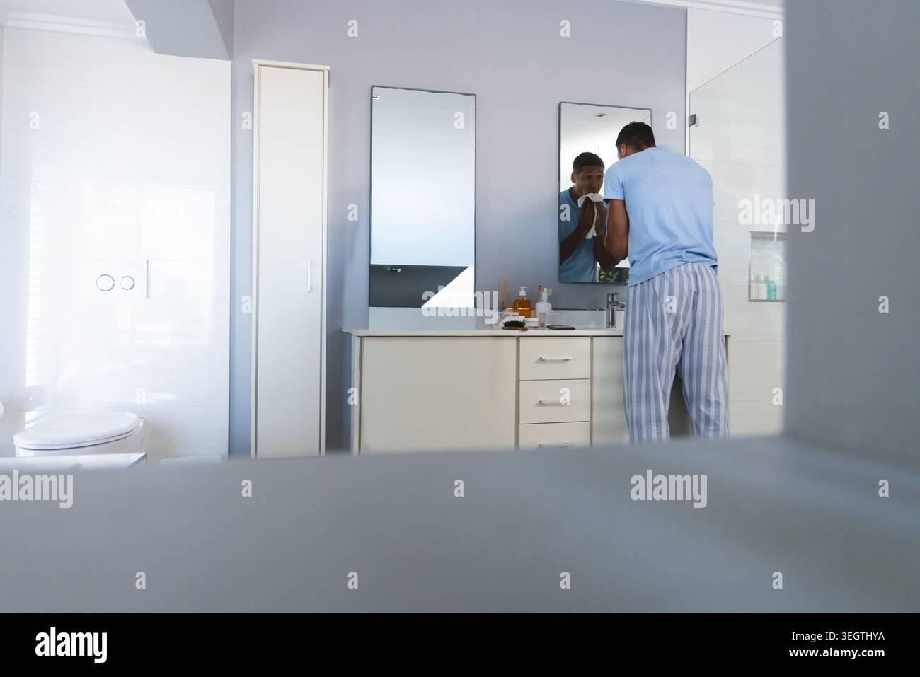 Male standing at bathroom vanity, wiping face with towel, in light blue tee, copy space Stock Photo
