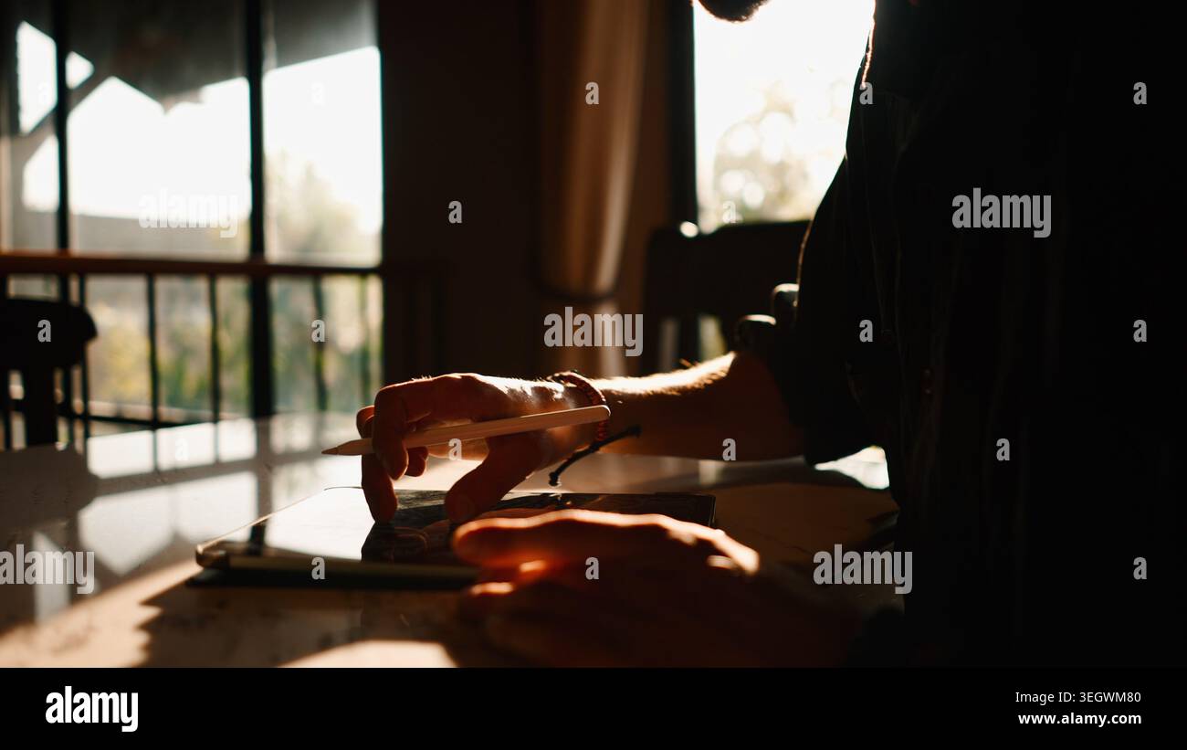 Man creating digital art on tablet using stylus, focusing on work at sunset Stock Photo