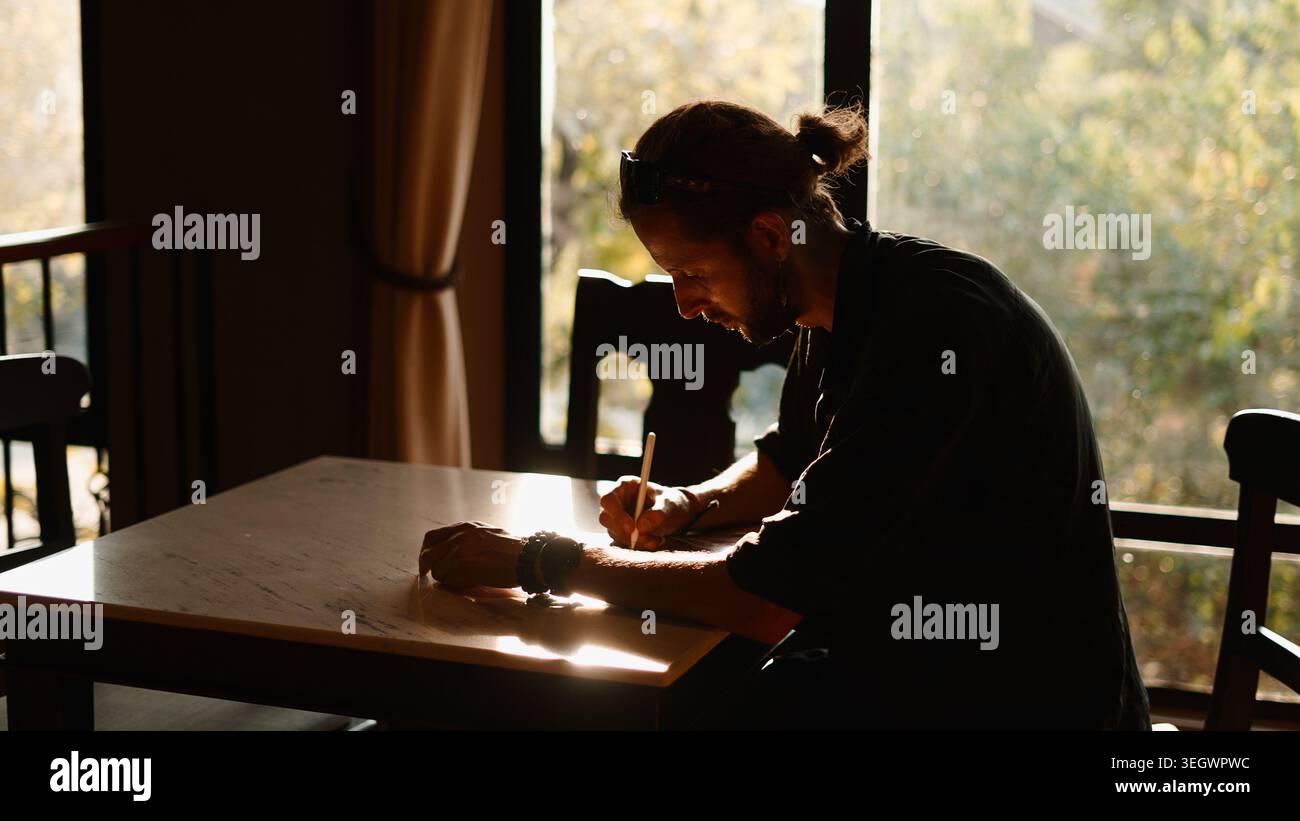 Man concentrating on writing or drawing at a table in warm sunlight ...