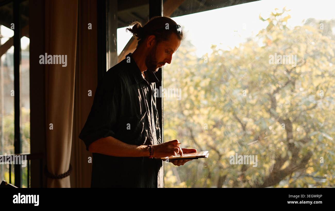 Creative man drawing on tablet near window, reflecting and working from home Stock Photo
