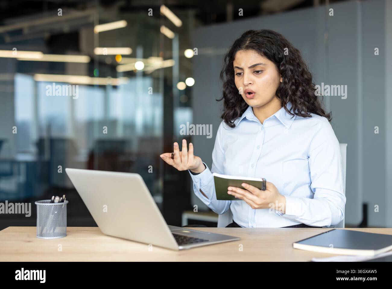 Young indian businesswoman using a laptop for an online video call in her office, feeling stressed and confused about potential technical problems while holding a notebook Stock Photo