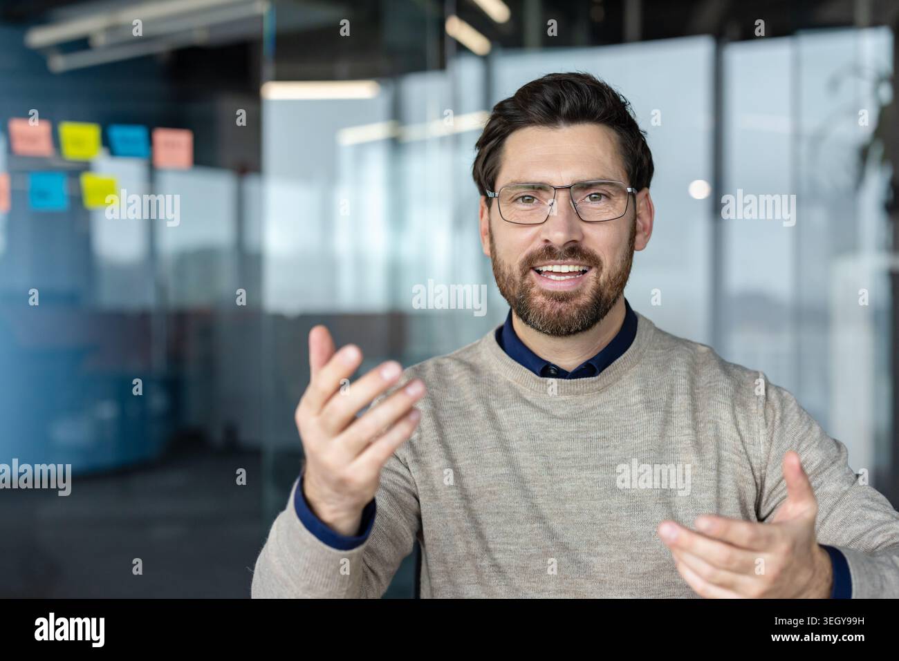 Professional man wearing glasses and a beard smiling while explaining a complex concept using hand gestures during a business meeting or online video call Stock Photo