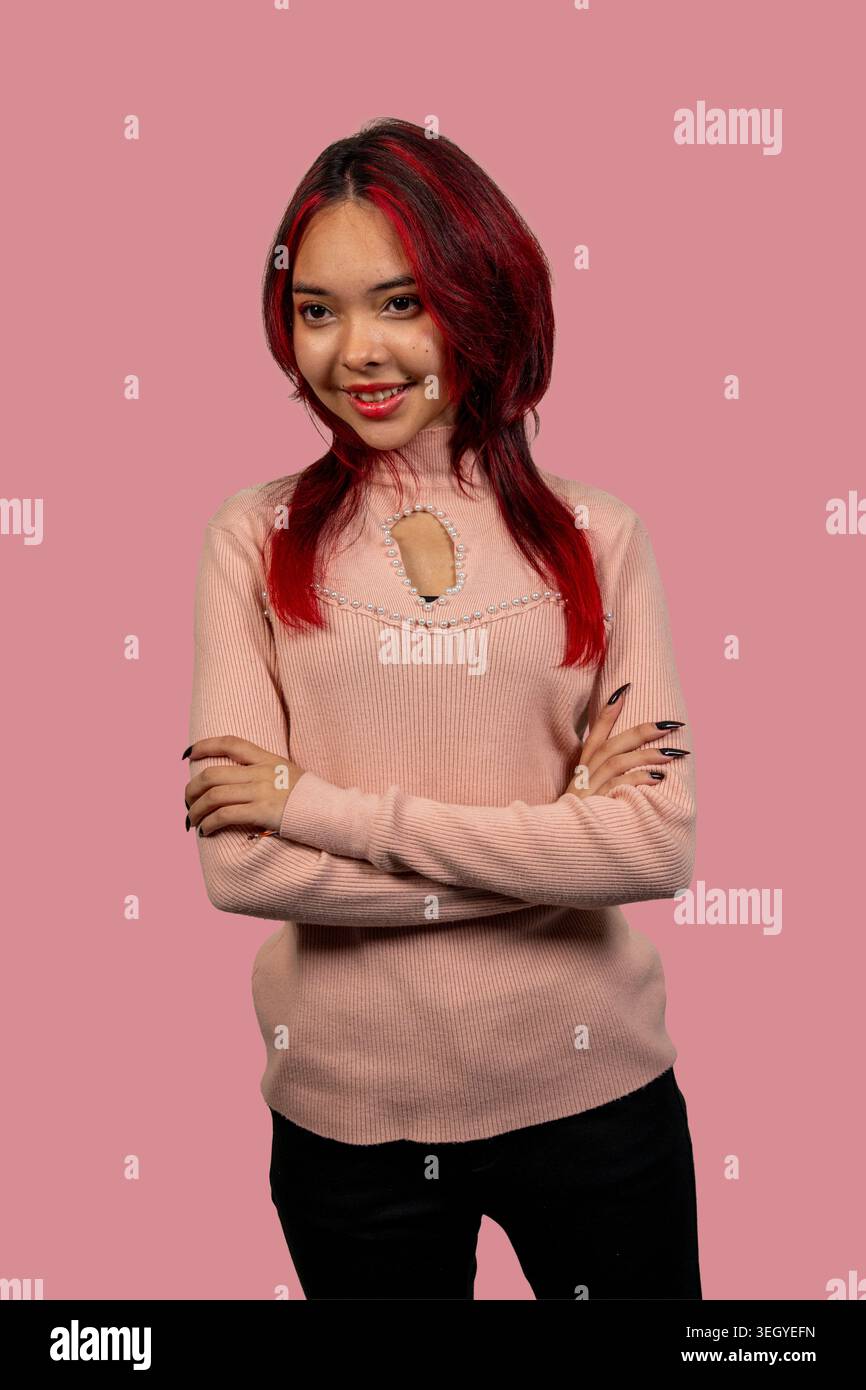 Young woman smiling while standing with her arms crossed on a pink studio background Stock Photo